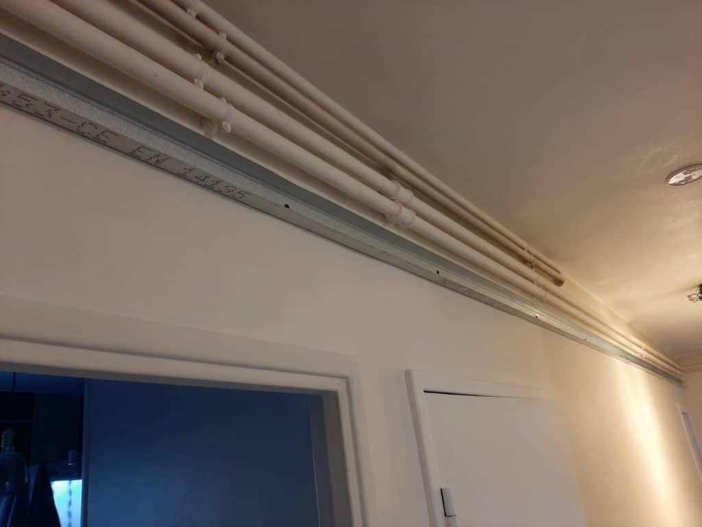 Des tuyaux blancs longeant le plafond au-dessus d'un rail métallique gravé '3S3-CE EN 14195' sur un mur clair.