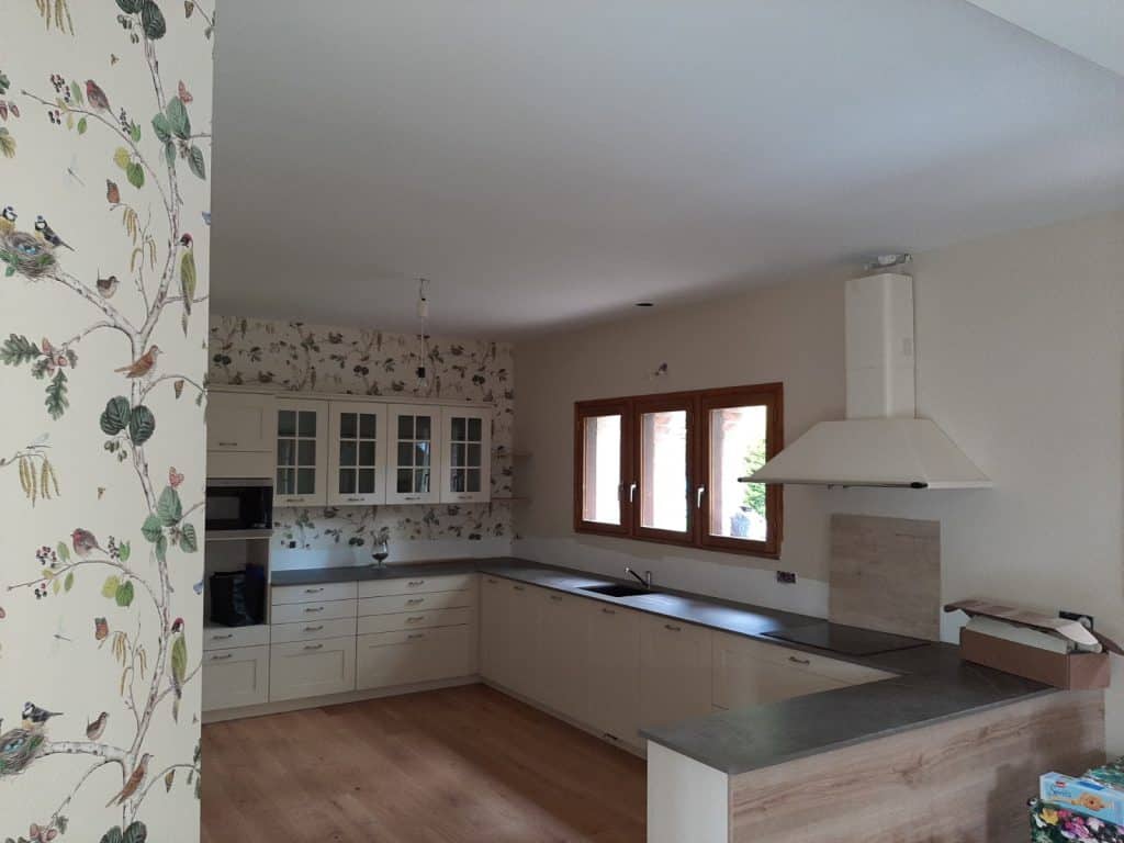 Cuisine moderne beige avec îlot, armoires claires, plan de travail foncé et hotte. Mur en papier peint à motif oiseaux et branchages, sol en bois clair.