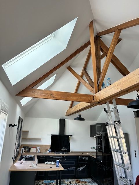 Cuisine mansardée moderne avec poutres en bois apparentes et deux velux. Mobilier noir, plan de travail clair et sol à motifs. Échelle.