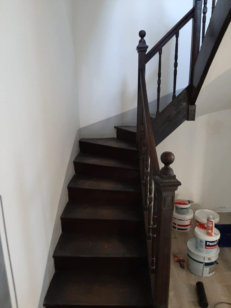 Escalier en bois sombre avec main courante et balustres, murs blancs immaculés. Des pots de peinture (PROG, MIN) et outils de rénovation sont au sol.
