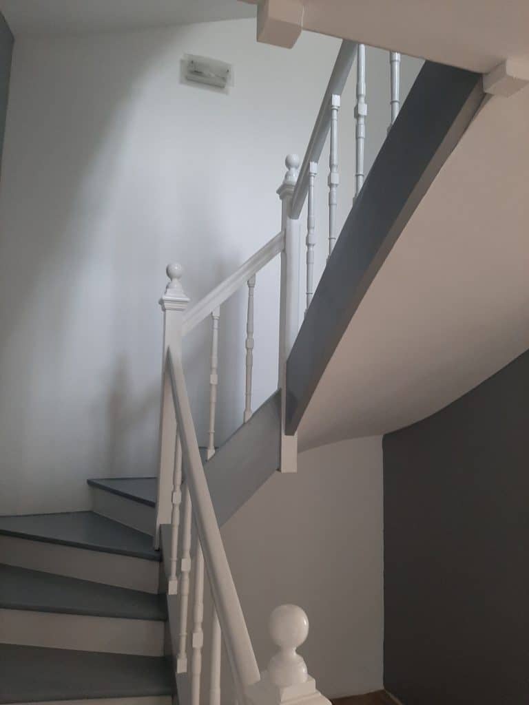Escalier bicolore moderne avec marches et sous-face grises, rampe blanche et murs clairs. Un luminaire mural est visible.