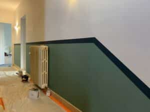 Couloir en rénovation avec murs bicolores vert, blanc et bande noire diagonale. Radiateur vintage. Pots de peinture "blime" au sol protégé.