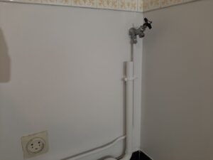 Coin de mur blanc, carrelage doré en haut. Robinet mural chromé à poignée noire et tuyaux blancs. Prise électrique jaunie en bas à gauche.
