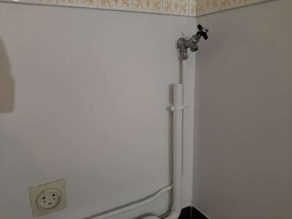 Coin de mur blanc, carrelage doré en haut. Robinet mural chromé à poignée noire et tuyaux blancs. Prise électrique jaunie en bas à gauche.